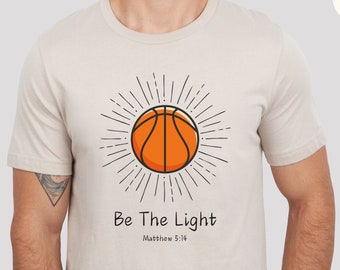 May include: A beige t-shirt with a basketball graphic and the text "Be The Light" with the scripture reference "Matthew 5:14".