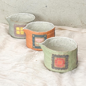 May include: Three handmade ceramic jugs in pastel shades. Each jug has a speckled interior and a decorative square design on the front. The colours include light green, orange, and a pale green with a yellow flower design.