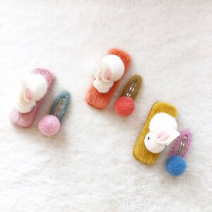 Könnte beinhalten: Drei Sätze Haarklammern mit einem flauschigen weißen Hasen-Anhänger, jede mit einer anderen farbigen Basis: rosa, orange und gelb. Jede Klammer hat auch einen kleinen, flauschigen Bommel in einer passenden Farbe.
