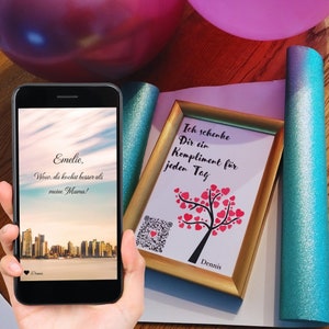 May include: A framed print with a gold frame and a white background. The print features a tree with red hearts for leaves and the text "Ich schenke Dir ein Kompliment für jeden Tag" and "Dennis". A smartphone with a black case is in the foreground, displaying a message that reads "Emelie, Wow, du kochst besser als meine Mama!" and a QR code.