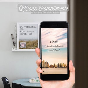 Op de afbeelding: Een zwarte smartphone met een QR-code met de tekst "Qr Code Komplimente" en "VERSCHENKE SCHONE WORTE". De telefoon toont ook een bericht met de tekst "Emelie, Wow, du kochst besser als meine Mama!"
