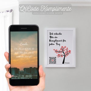 May include: A white framed print with a pink tree with hearts on the branches. The text on the print reads "Ich schenke Dir ein Kompliment für jeden Tag" and "Dennis". A QR code is in the bottom left corner of the print. A smartphone is in the bottom left corner of the image, displaying a message that reads "Emelie, Die Art, wie du lachst, dir an die Nase fasst, wenn du nachdenkst, ist so süß!"