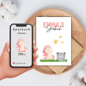 Könnte beinhalten: Ein Smartphone zeigt eine Sparbuch-Grafik mit einer Einhorn-Illustration und dem Text "Sparbuch - Emma - 250 EUR". Eine passende Karte mit "EMMA'S Sparbuch" und einer Einhorn-Illustration ist ebenfalls abgebildet.