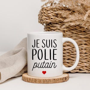 Peut inclure: Un mug en céramique blanche avec le texte imprimé en noir "JE SUIS POLIE putain" et une petite icône de cœur rouge centrée sous le texte. Le mug possède une anse standard et repose sur un dessous de verre en bois à bord écorcé.