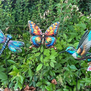 May include: Three colorful metal garden stakes: a dragonfly, a butterfly, and a hummingbird. The dragonfly is blue and teal, the butterfly has orange, yellow, and blue wings, and the hummingbird is green, blue, and orange.