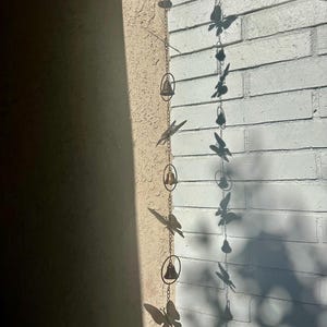 May include: A decorative metal wind chime featuring butterfly and bell accents. The wind chime is suspended from a chain and includes several butterfly cutouts and small bells. The color is a bronze-like hue, and it hangs against a textured wall.