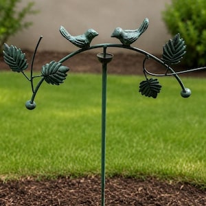 May include: A green metal garden stake featuring a central vertical rod topped with an arched branch. Two small birds face each other on the arch, surrounded by detailed metal leaves and small spherical accents on the ends of the branches.