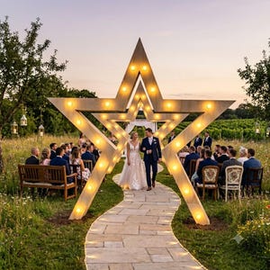 Op de afbeelding: Een grote, lichtgekleurde houten boog in de vorm van een ster, verlicht met warm gloeiende lampen. De structuur staat over een stenen pad in een grasveld en dient als achtergrond voor een huwelijksceremonie met gasten op houten banken.