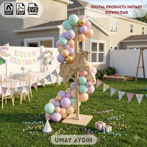 May include: A wooden carousel horse cutout mounted on a vertical stand, decorated with a balloon arch in pastel shades of pink, blue, purple, and gold. The display sits on a grassy lawn with party decorations, including a banner and gift boxes.