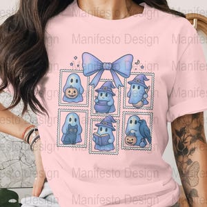 Puede incluir: Camiseta rosa claro con un diseño de Halloween. El diseño incluye personajes de fantasmas azules en marcos de sellos postales, un gran lazo azul y el texto "Manifesto Design". Los fantasmas sostienen calabazas, usan sombreros de bruja y llevan un gato negro.