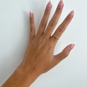 Op de afbeelding: Een gouden ring met een klein kettinkje en drie gekleurde edelstenen. De ring zit aan een vinger met witte french manicure nagels.