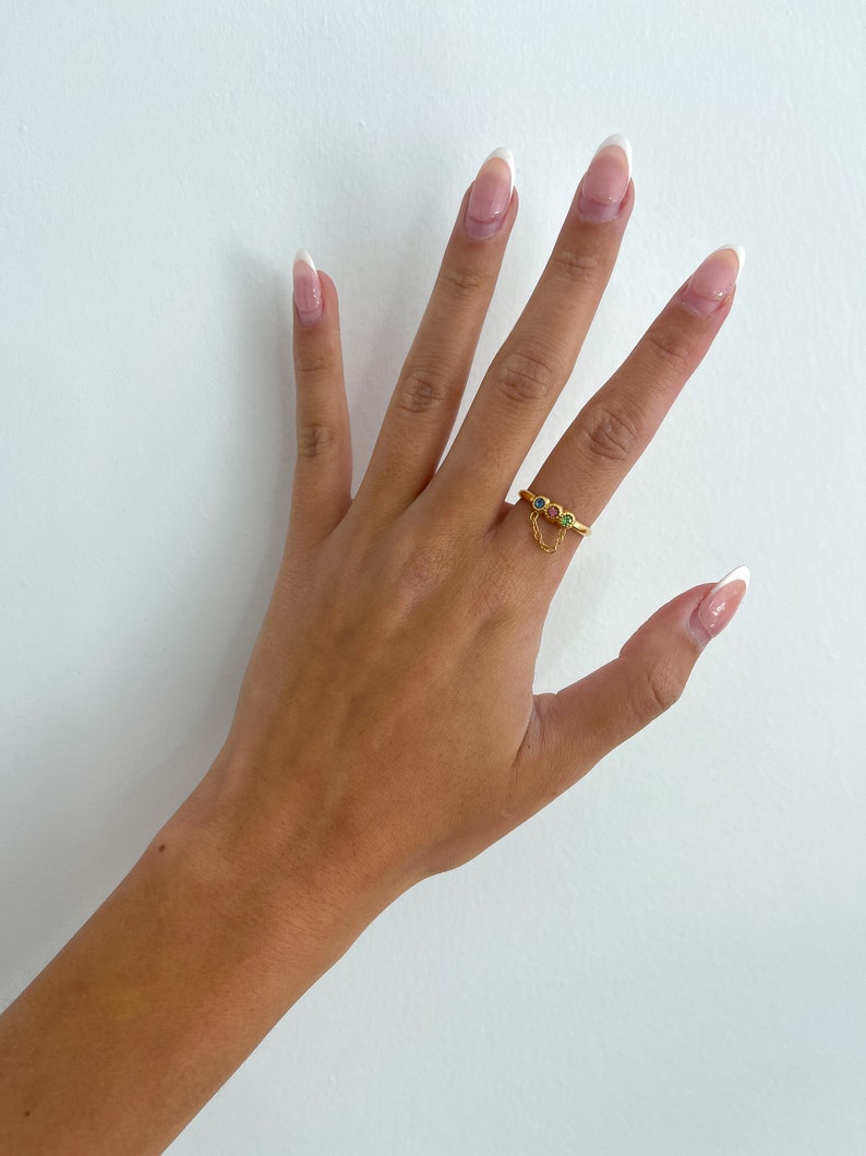 Op de afbeelding: Een gouden ring met een klein kettinkje en drie gekleurde edelstenen. De ring zit aan een vinger met witte french manicure nagels.