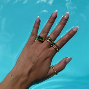 May include: A hand with white nail polish is wearing several gold rings, including one with a green stone. The hand is resting on a blue surface.