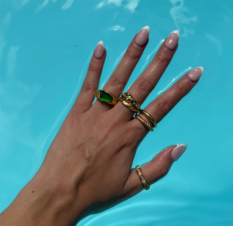 May include: A hand with white nail polish is wearing several gold rings, including one with a green stone. The hand is resting on a blue surface.