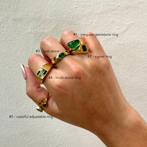 May include: A hand wearing five gold rings with green gemstones. The rings are labeled #1 - irregular gemstone ring, #2 - signet ring, #3 - oval stone, #4 - multi-stone ring, and #5 - colorful adjustable ring.
