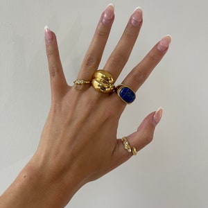 Op de afbeelding: Een hand met vier gouden ringen. De ringen zijn een dikke gouden band, een ring met een blauwe steen, een ring met een klein kettinkje en een ring met kleine heldere steentjes. De nagels zijn gelakt met een witte tip manicure.