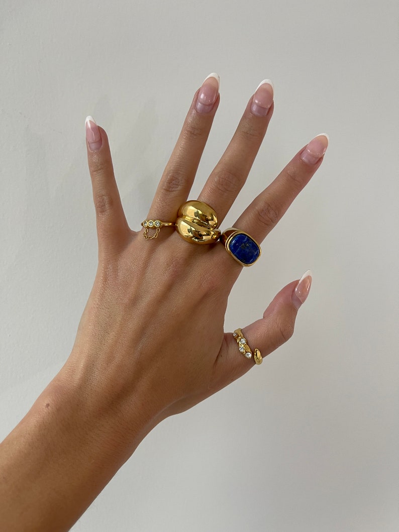 Op de afbeelding: Een hand met vier gouden ringen. De ringen zijn een dikke gouden band, een ring met een blauwe steen, een ring met een klein kettinkje en een ring met kleine heldere steentjes. De nagels zijn gelakt met een witte tip manicure.
