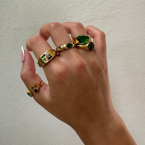 May include: A hand wearing four gold rings with green gemstones. The rings are stacked on two fingers.
