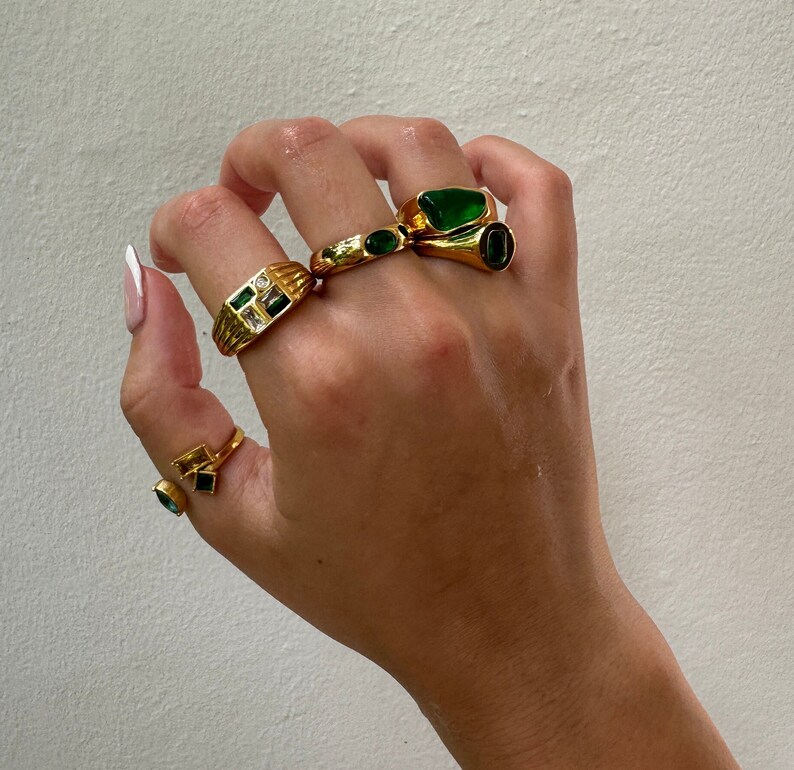 May include: A hand wearing four gold rings with green gemstones. The rings are stacked on two fingers.