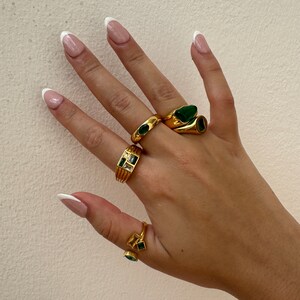 May include: A hand wearing four gold rings with green gemstones. The rings are stacked on two fingers. The nails are painted with a white french manicure.