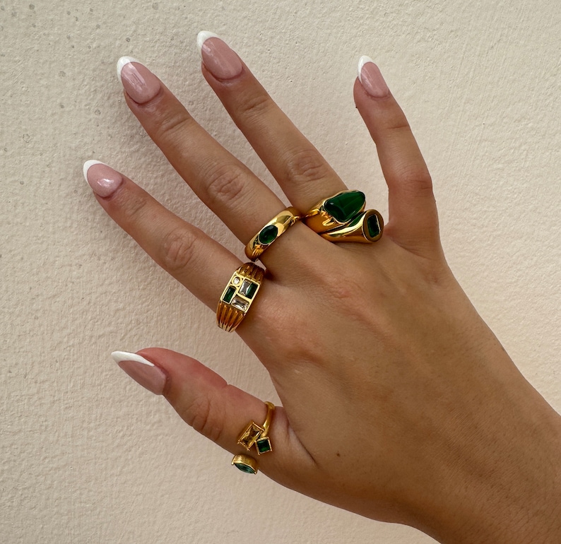 May include: A hand wearing four gold rings with green gemstones. The rings are stacked on two fingers. The nails are painted with a white french manicure.
