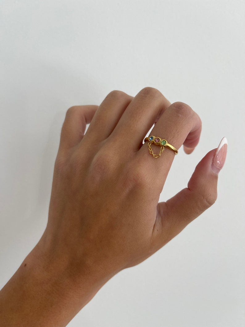 Op de afbeelding: Gouden ring met een hangende ketting en drie gekleurde edelstenen. De ring zit aan een vinger.