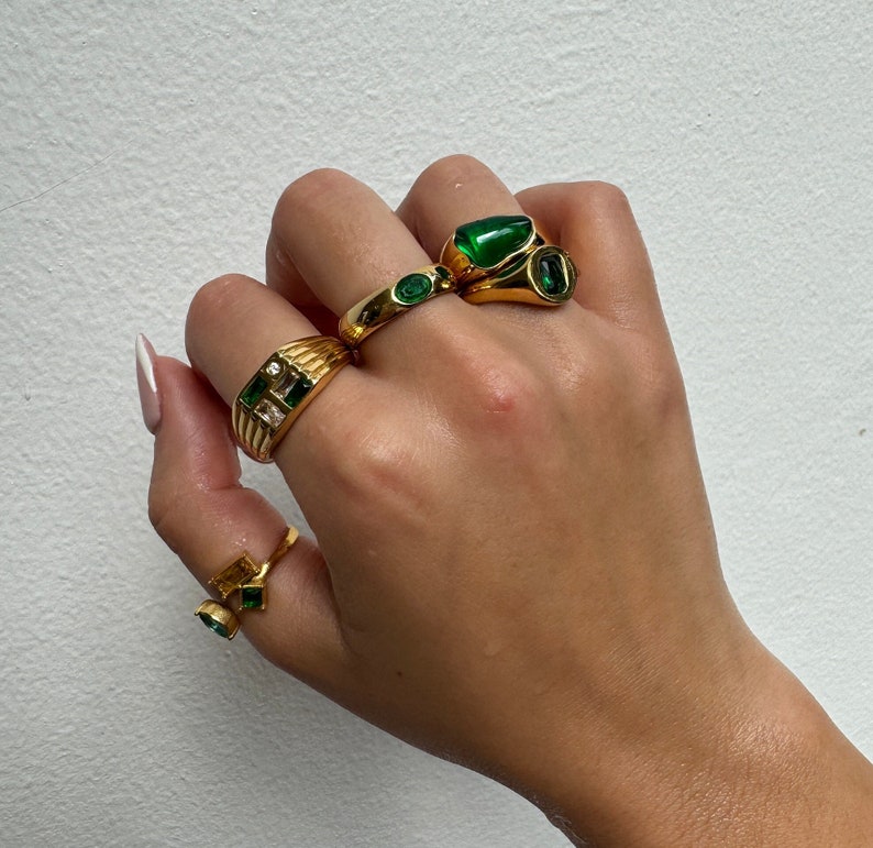 May include: A hand wearing four gold rings with green gemstones. The rings are stacked on two fingers.