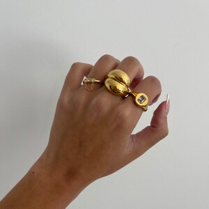 Op de afbeelding: Een hand met drie gouden ringen. De ring op de wijsvinger is een grote, ronde, gouden ring. De ring op de middelvinger is een kleinere, vierkante ring met een heldere steen. De ring op de pink is een dunne gouden band met een kleine ketting.