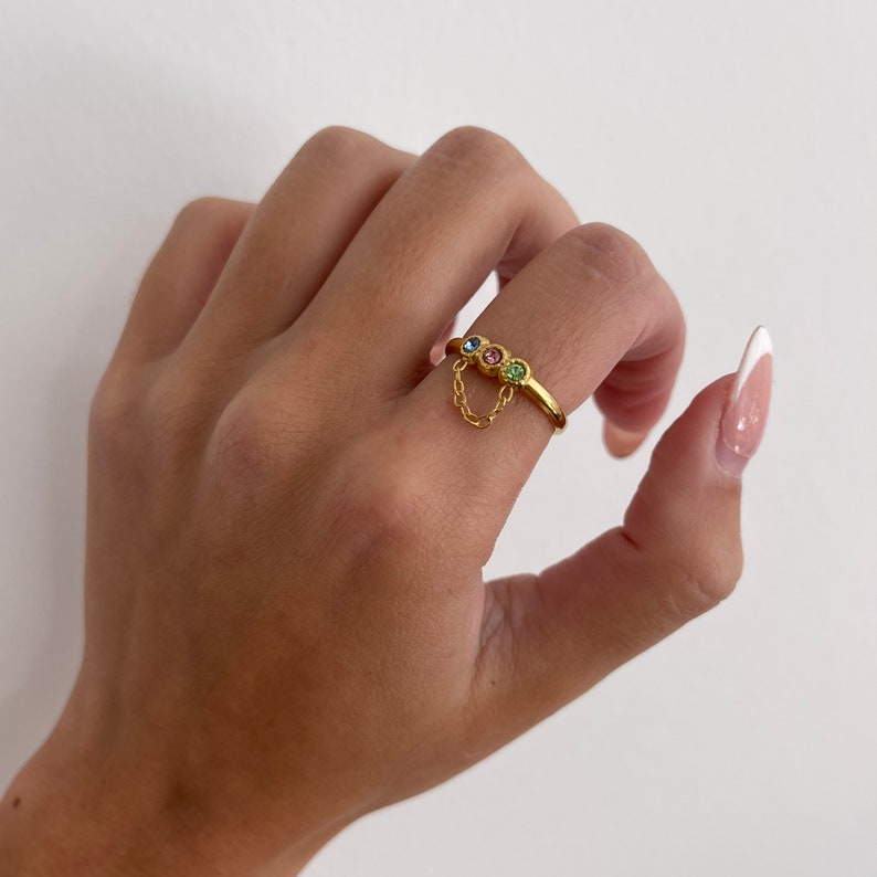 Op de afbeelding: Gouden ring met een hangende ketting en drie gekleurde edelstenen. De edelstenen zijn blauw, roze en groen.