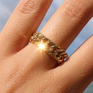May include: A gold chain ring with a thick, chunky design. The ring is worn on a finger and is made of a shiny metal.