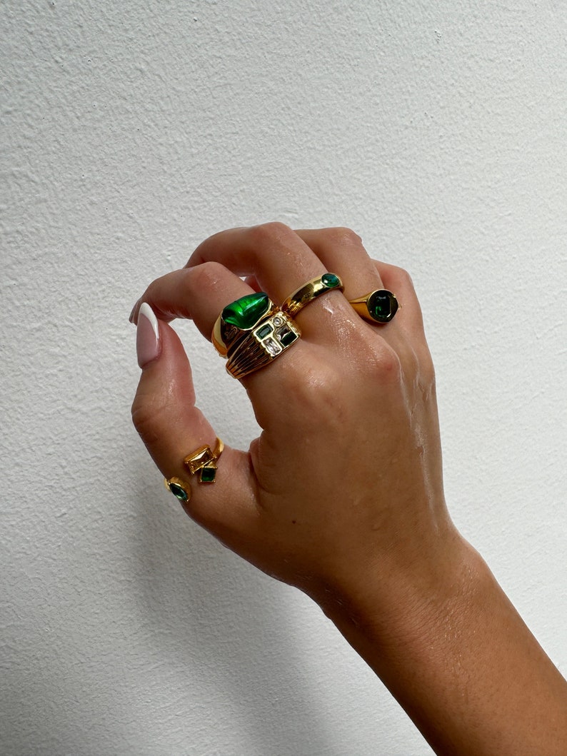 May include: A hand wearing four gold rings with green gemstones. The rings are stacked on the middle finger and the index finger. The rings are all different styles and sizes.