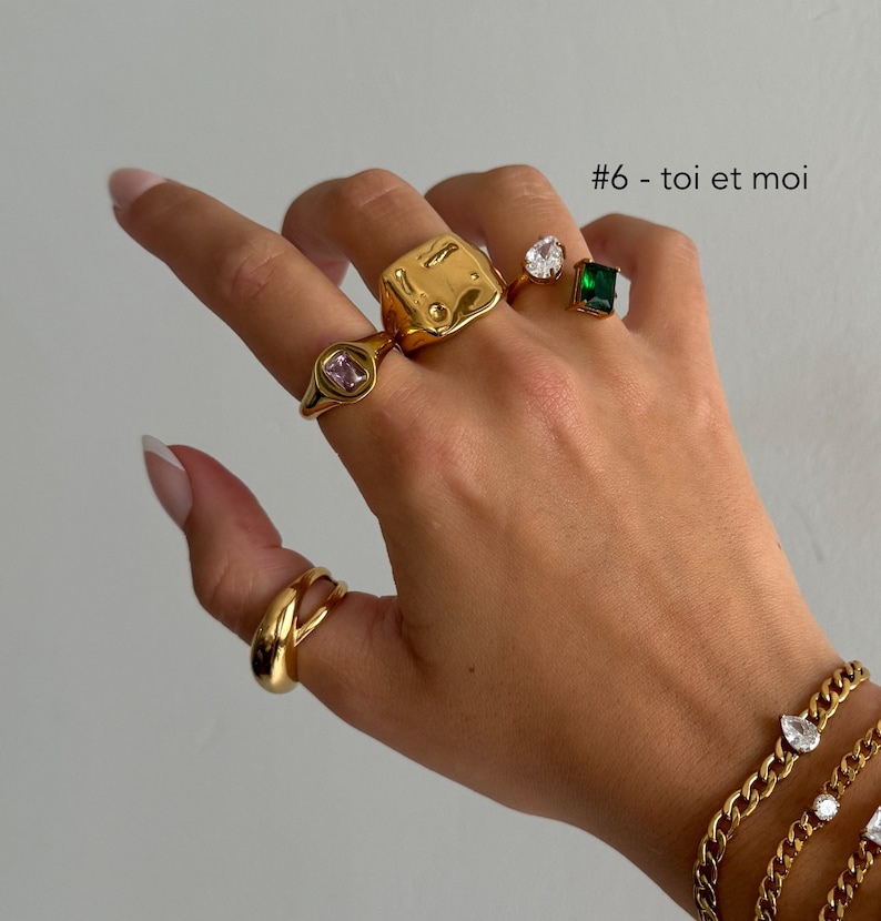 May include: A hand wearing a set of gold rings with various gemstones, including a pink square stone, a clear round stone, and a green square stone. The hand is also wearing a gold chain bracelet with a clear teardrop-shaped stone. The text '#6 - toi et moi' is visible in the image.