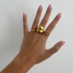 Op de afbeelding: Een hand met drie gouden ringen. De ringen zijn allemaal in verschillende stijlen. Een ring is een dikke band met een afgeronde vorm. Een andere ring is een dunne band met een klein kettinkje en drie kleine steentjes. De derde ring is een dunne band met een grote vierkante steen.