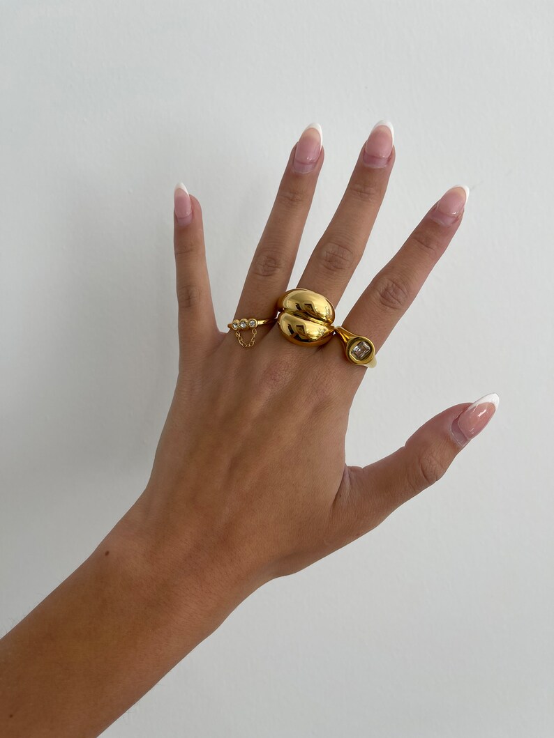 Op de afbeelding: Een hand met drie gouden ringen. De ringen zijn allemaal in verschillende stijlen. Een ring is een dikke band met een afgeronde vorm. Een andere ring is een dunne band met een klein kettinkje en drie kleine steentjes. De derde ring is een dunne band met een grote vierkante steen.