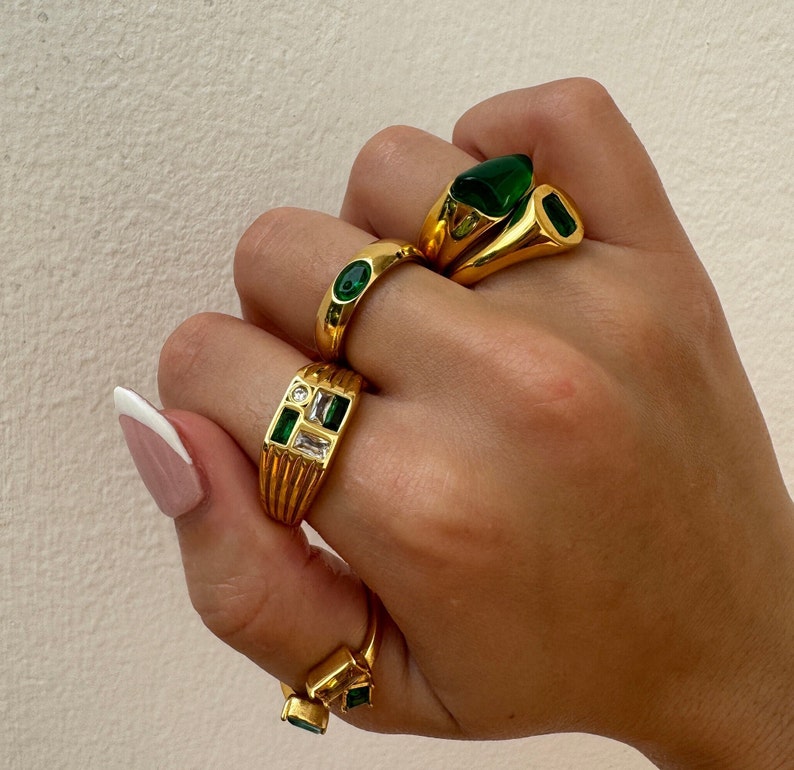 May include: A hand wearing four gold rings with green and clear gemstones. The rings are stacked on each other and are a statement piece of jewelry.