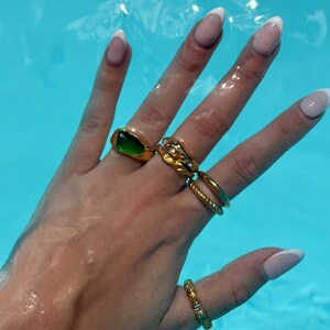 May include: A hand wearing a set of gold rings, including a green stone ring, a ring with a textured design, and a thin band ring. The hand is also wearing a gold ring on the pinky finger.