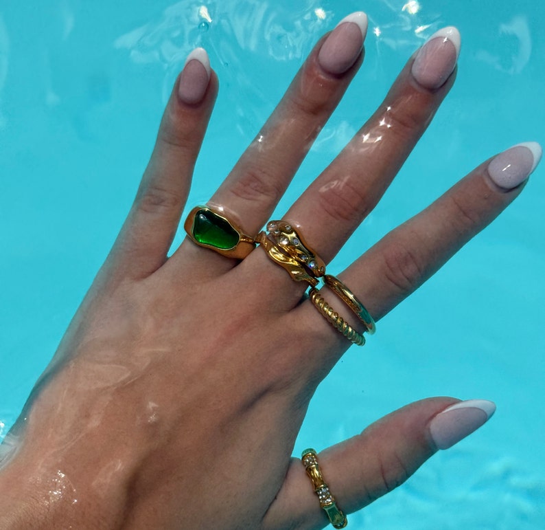 May include: A hand wearing a set of gold rings, including a green stone ring, a ring with a textured design, and a thin band ring. The hand is also wearing a gold ring on the pinky finger.
