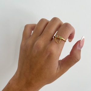 Op de afbeelding: Gouden ring met een hangende ketting en drie gekleurde edelstenen. De edelstenen zijn roze, blauw en groen.