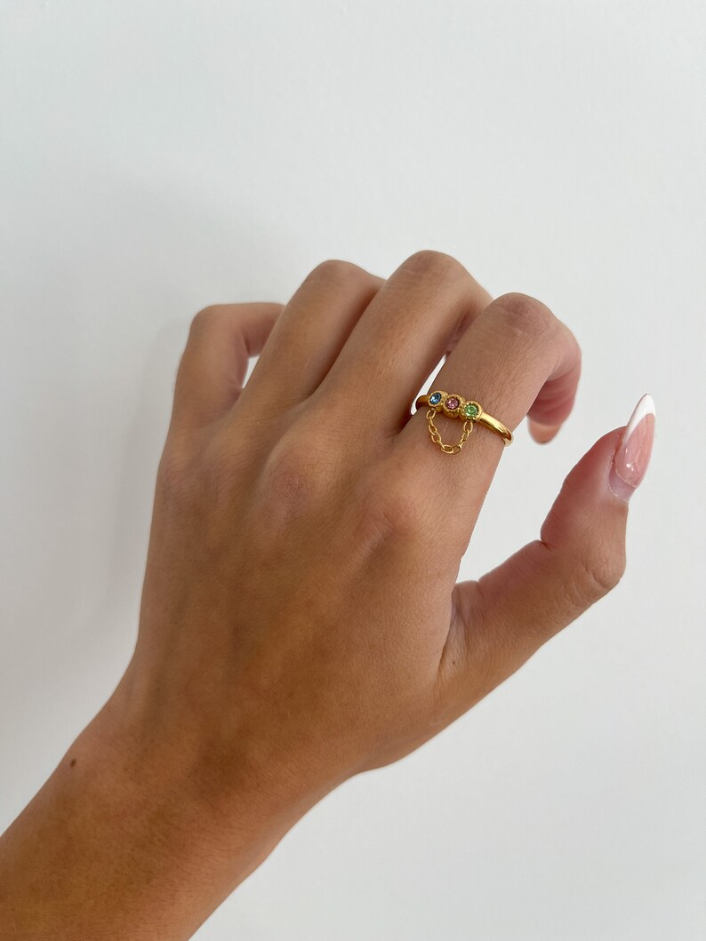 Op de afbeelding: Gouden ring met een hangende ketting en drie gekleurde edelstenen. De edelstenen zijn roze, blauw en groen.