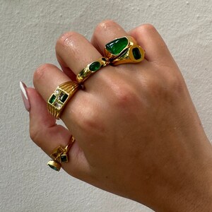 May include: A hand wearing four gold rings with green and clear gemstones. The rings are stacked on two fingers.