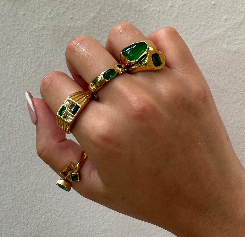 May include: A hand wearing four gold rings with green and clear gemstones. The rings are stacked on two fingers.