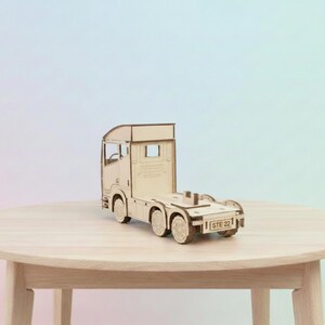 May include: A detailed wooden model of a truck cab and chassis, with visible wheels and design features. The model is made of light-coloured wood and is placed on a round wooden table. The text "STE 22" is visible on the back.