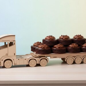 May include: A wooden toy lorry transporting chocolate cupcakes with chocolate frosting. The cupcakes are stacked on the flatbed. The lorry is constructed from light-coloured wood and has detailed features.