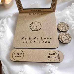 May include: A wooden wedding game with a soccer theme. The game features a wooden goal, a soccer ball, and two smaller soccer balls. The game is personalized with the text "Mr. & Mr. Love 17.08.2024".