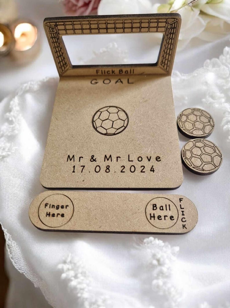 May include: A wooden wedding game with a soccer theme. The game features a wooden goal, a soccer ball, and two smaller soccer balls. The game is personalized with the text "Mr. & Mr. Love 17.08.2024".