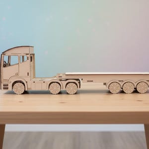 May include: A wooden toy articulated lorry and trailer, constructed from light-coloured wood. The lorry has detailed cutouts for windows and wheels, and the trailer has a flatbed design. The toy is set on a wooden table against a soft, colourful background.