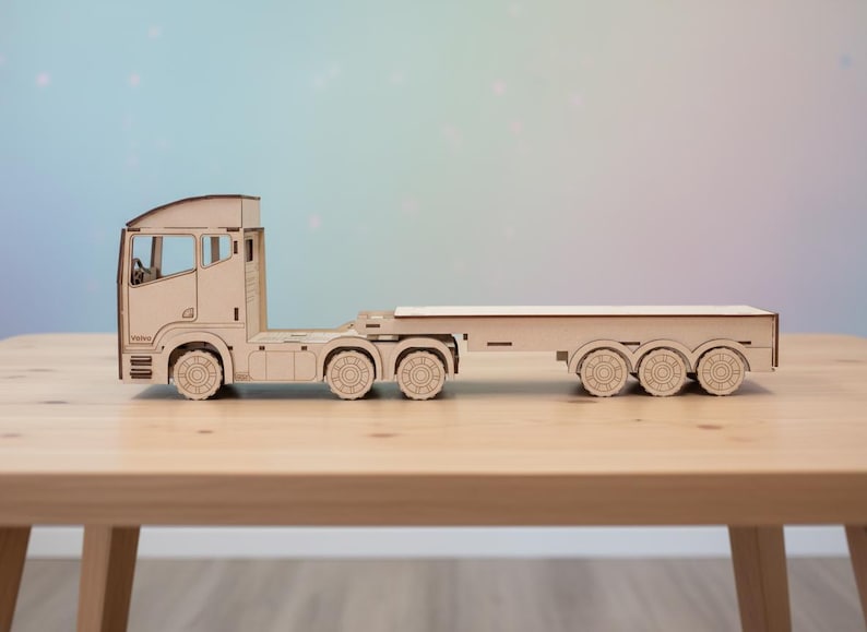 May include: A wooden toy articulated lorry and trailer, constructed from light-coloured wood. The lorry has detailed cutouts for windows and wheels, and the trailer has a flatbed design. The toy is set on a wooden table against a soft, colourful background.