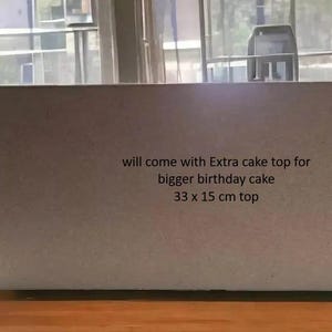 May include: A gray cardboard truck-shaped cake topper with text that reads "will come with Extra cake top for bigger birthday cake 33 x 15 cm top". The truck has wheels and a flatbed. The topper is on a wooden surface.