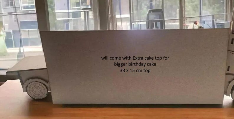 May include: A gray cardboard truck-shaped cake topper with text that reads "will come with Extra cake top for bigger birthday cake 33 x 15 cm top". The truck has wheels and a flatbed. The topper is on a wooden surface.