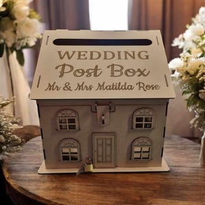 May include: A house-shaped wooden wedding post box. The box is engraved with the words "WEDDING Post Box" and "Mr & Mrs Matilda Rose". It has a slot for cards, arched windows, a door, and a small lock with a key. The box is made of wood.